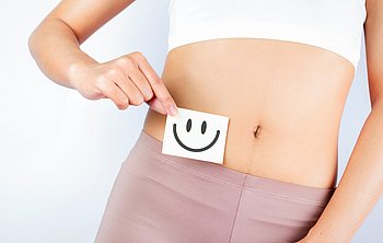 Hier sieht man eine Frau die einen Zettel mit einem lachenden Smiley vor ihren Bauch hält