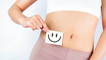 Hier sieht man eine Frau die einen Zettel mit einem lachenden Smiley vor ihren Bauch hält