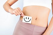 Hier sieht man eine Frau die einen Zettel mit einem lachenden Smiley vor ihren Bauch hält