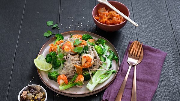 Rezept Buchweizennudeln Das fertig zubereitete Buchweizennudel-Gericht mit Garnelen, Pak Choi und Brokkoli auf einem Teller angerichtet. Daneben sieht man eine Schale mit Kimchi. © www.eatsmarter.de, Foto: Iris Lange-Fricke